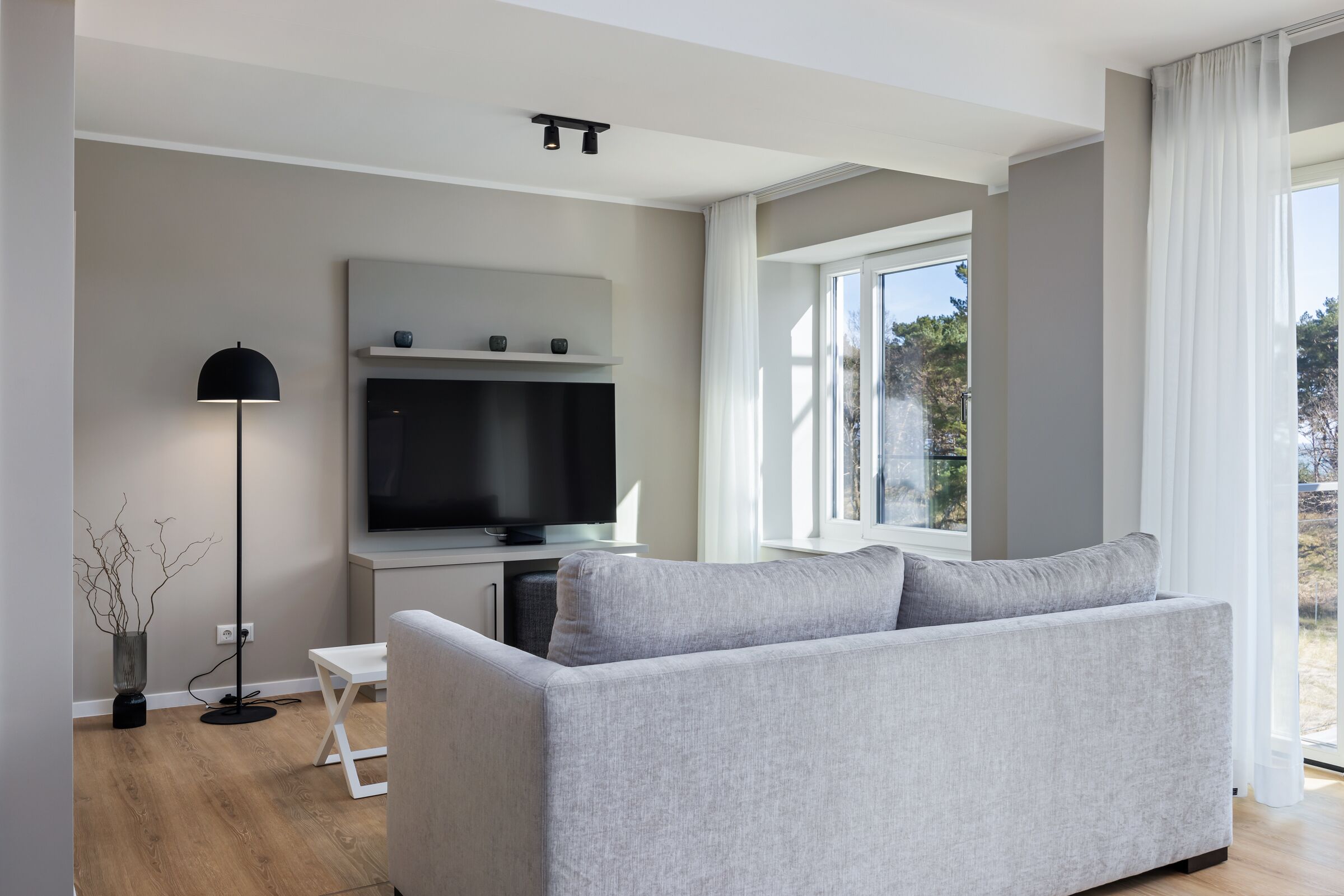 Un salón moderno con un sofá gris, un televisor de pantalla plana, una lámpara de pie y una ventana con vistas a árboles.