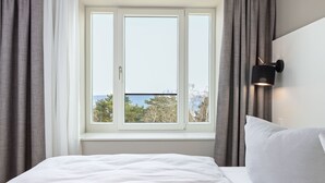 Quarto com uma grande janela, uma cama com roupa de cama branca, um travesseiro branco e luminária fixada na parede.