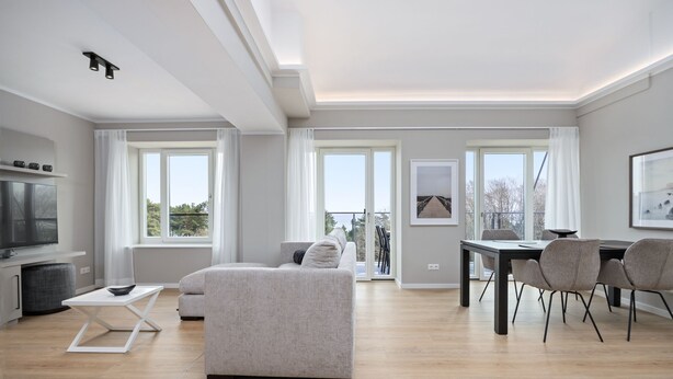 Uma sala de estar moderna com um sofá, uma mesa de jantar com cadeiras, uma televisão e janelas amplas.