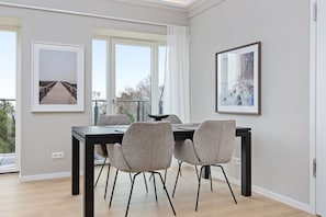 Un comedor moderno con una mesa negra y cuatro sillas grises, obras de arte enmarcadas en las paredes y ventanas amplias con vista a árboles.