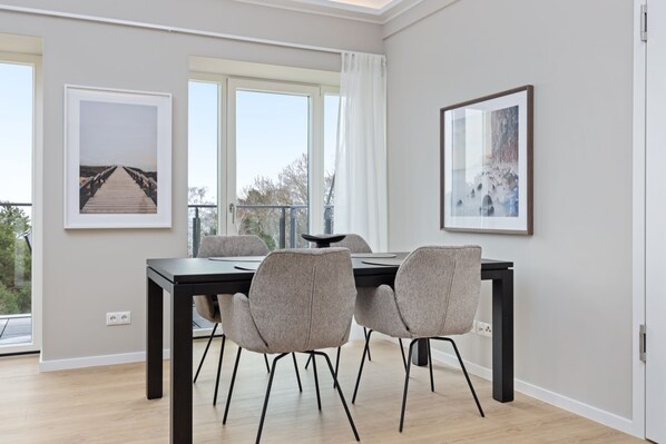 A modern dining area with a black table and four grey chairs, framed artwork on the walls, and large windows with a view of trees.