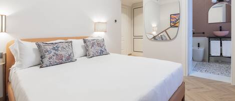 A bedroom with a large bed, a bedside table, a wall-mounted mirror, and a bathroom visible through an open door.