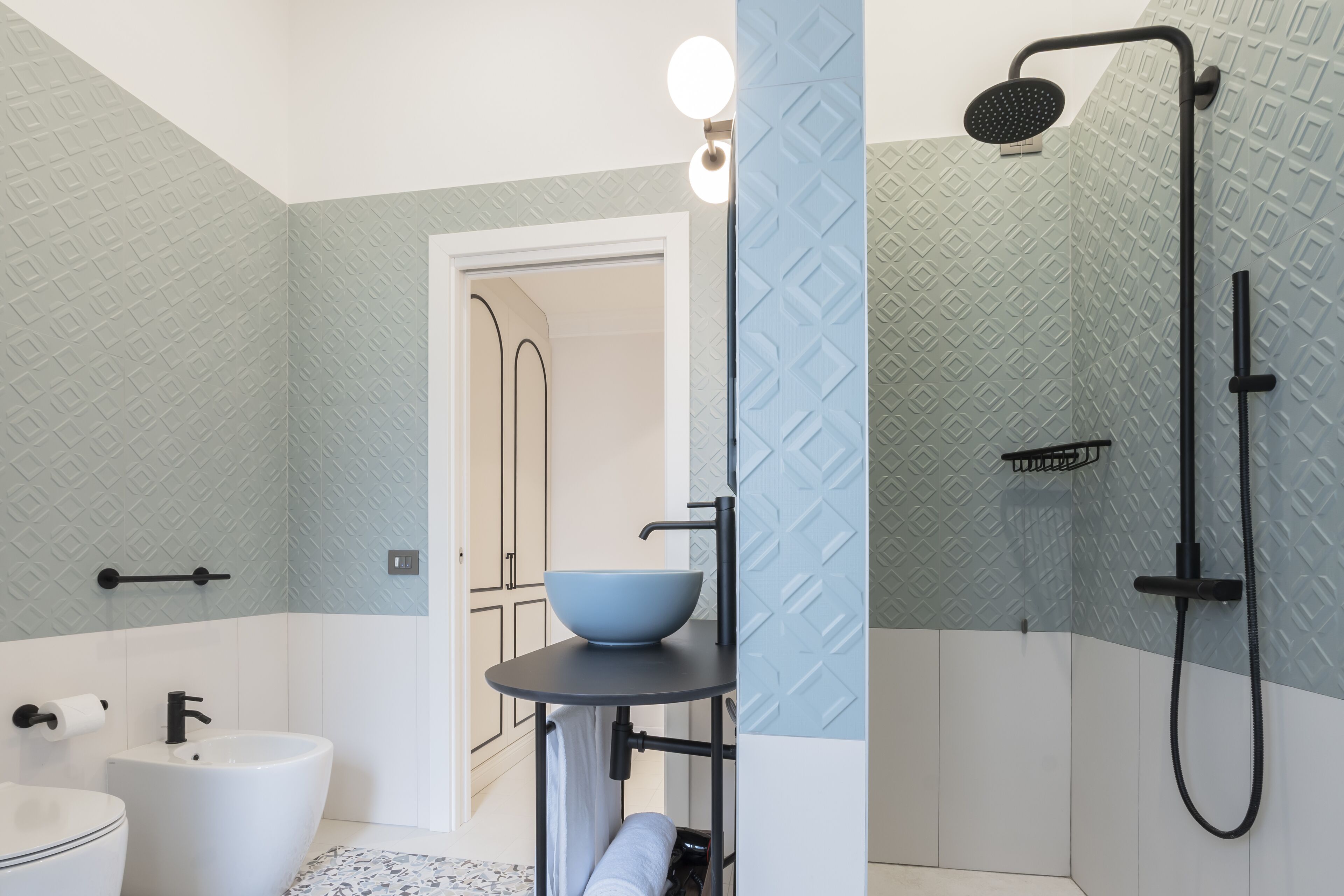 A bathroom with a white sink, a black faucet, a white toilet, a blue bowl on a black stand, and a black shower with a handheld showerhead.