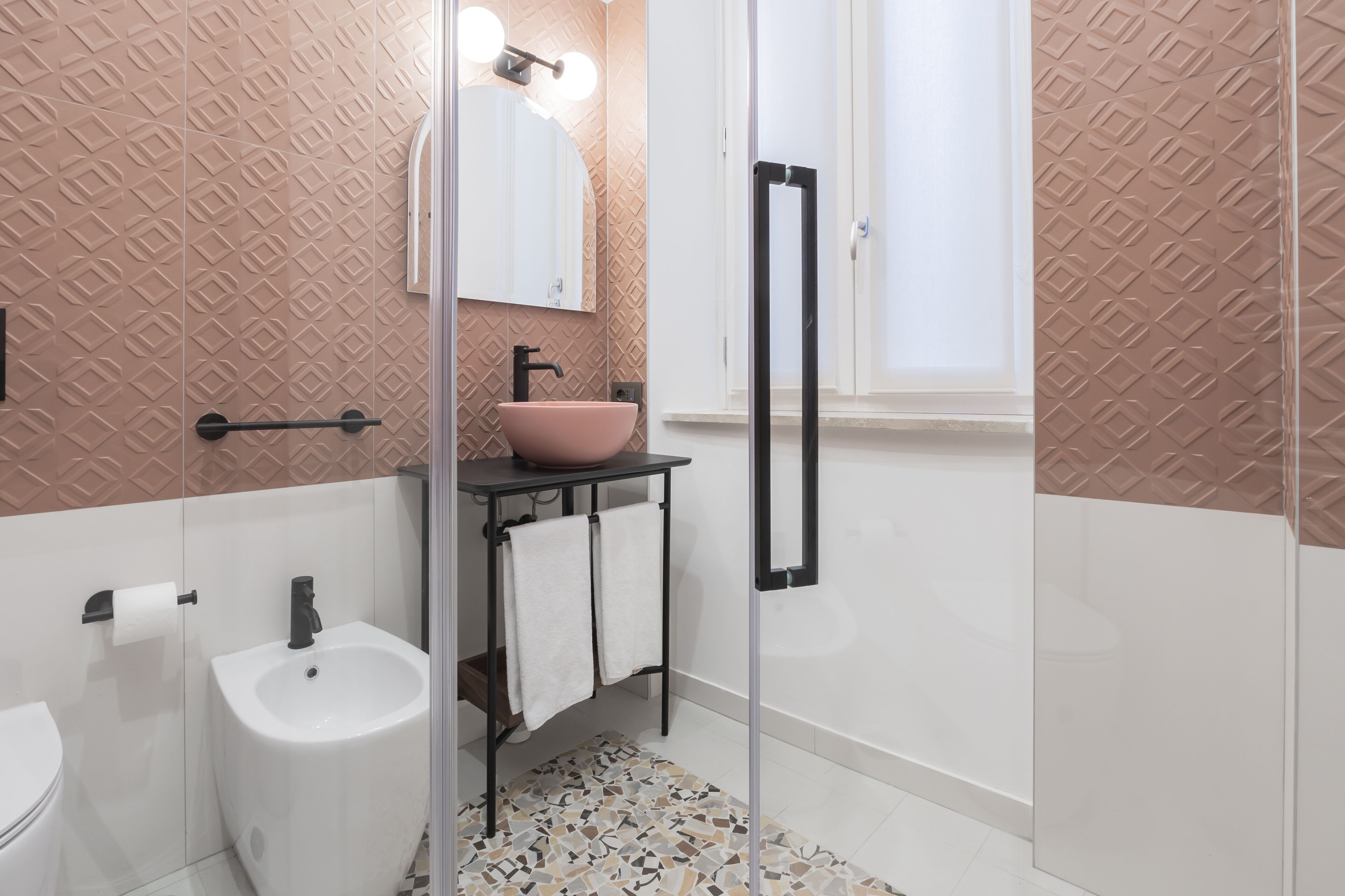 A bathroom with a sink, bidet, and toilet. There is a towel rack with towels, a mirror, and a shower door.
