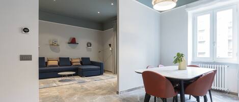 A modern living room with a blue sofa, a dining table with chairs, and a large window with a view.
