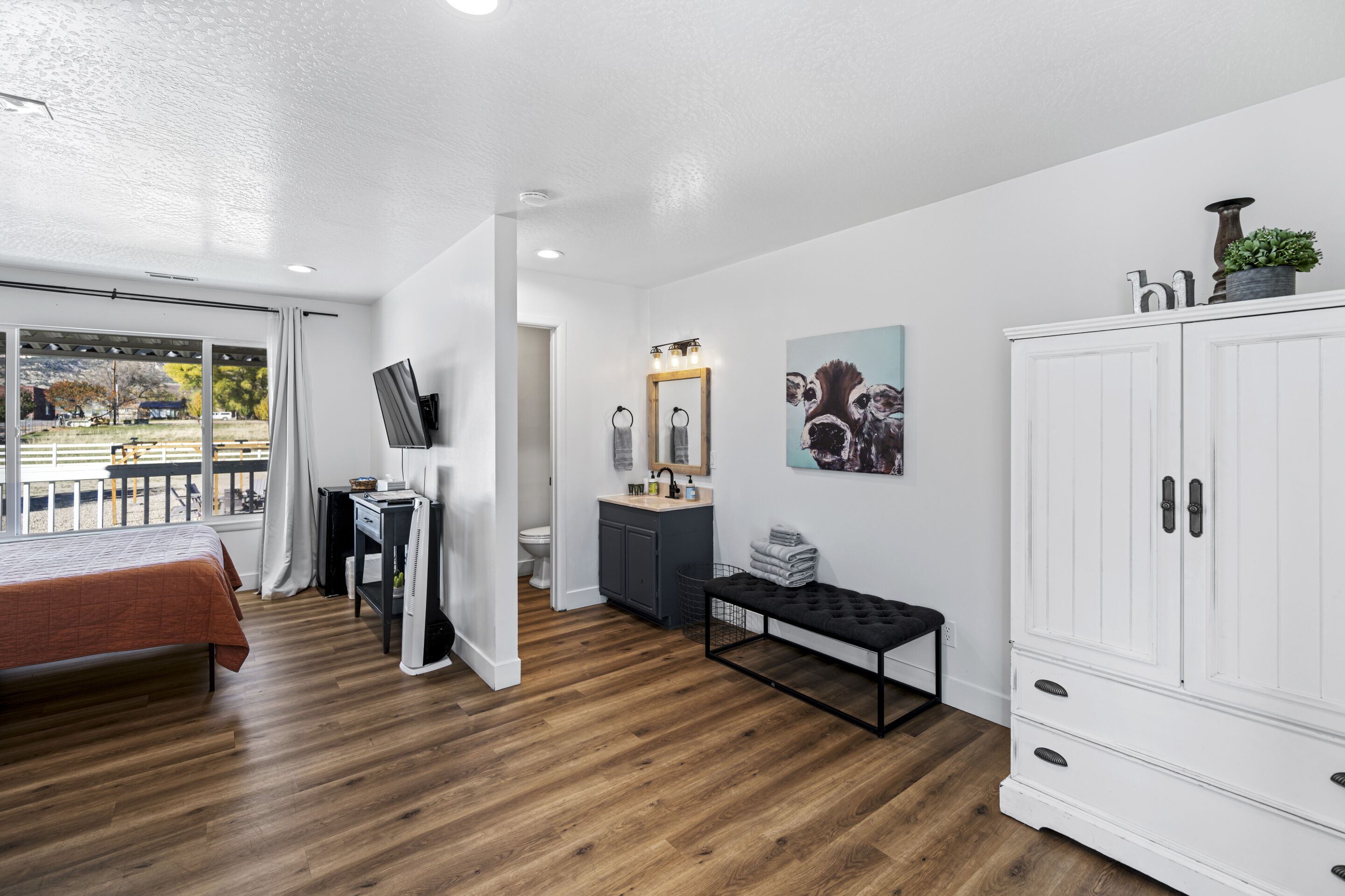 Una habitación moderna con cama, escritorio, banco, armario, baño con lavamanos y espejo, y obras de arte en las paredes.