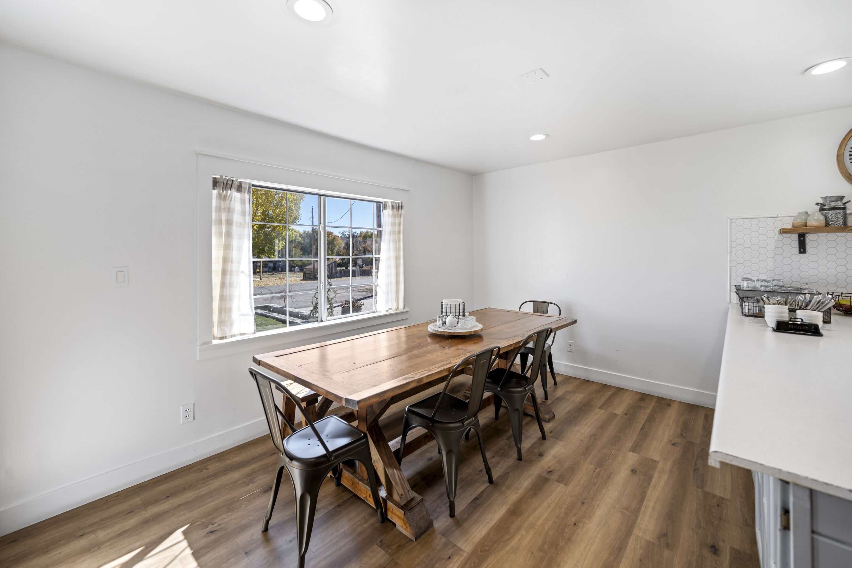 A dining area with a wooden table and chairs, a window with curtains, and a kitchen counter with utensils.