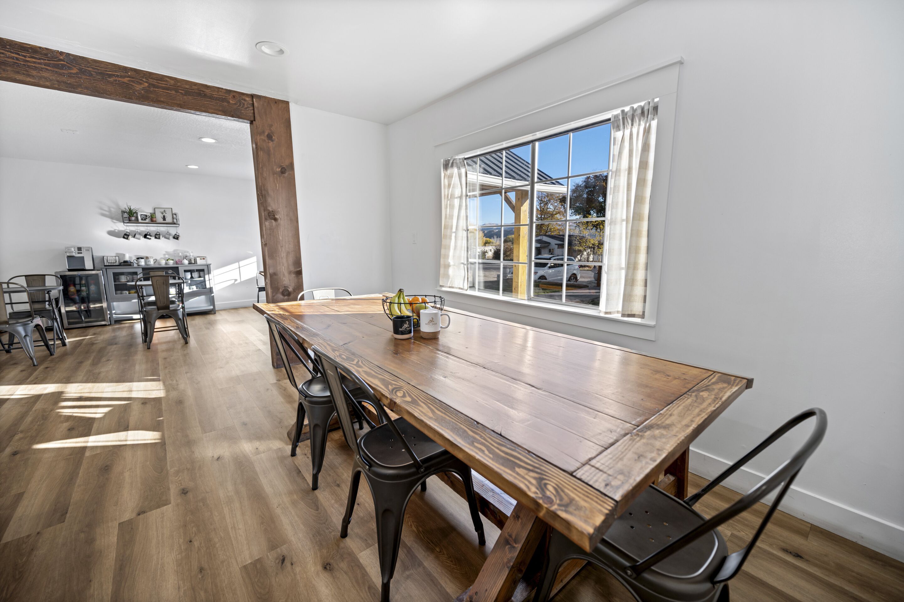 A spacious dining area with a long wooden table and modern chairs, a kitchen in the background, and a large window with curtains.