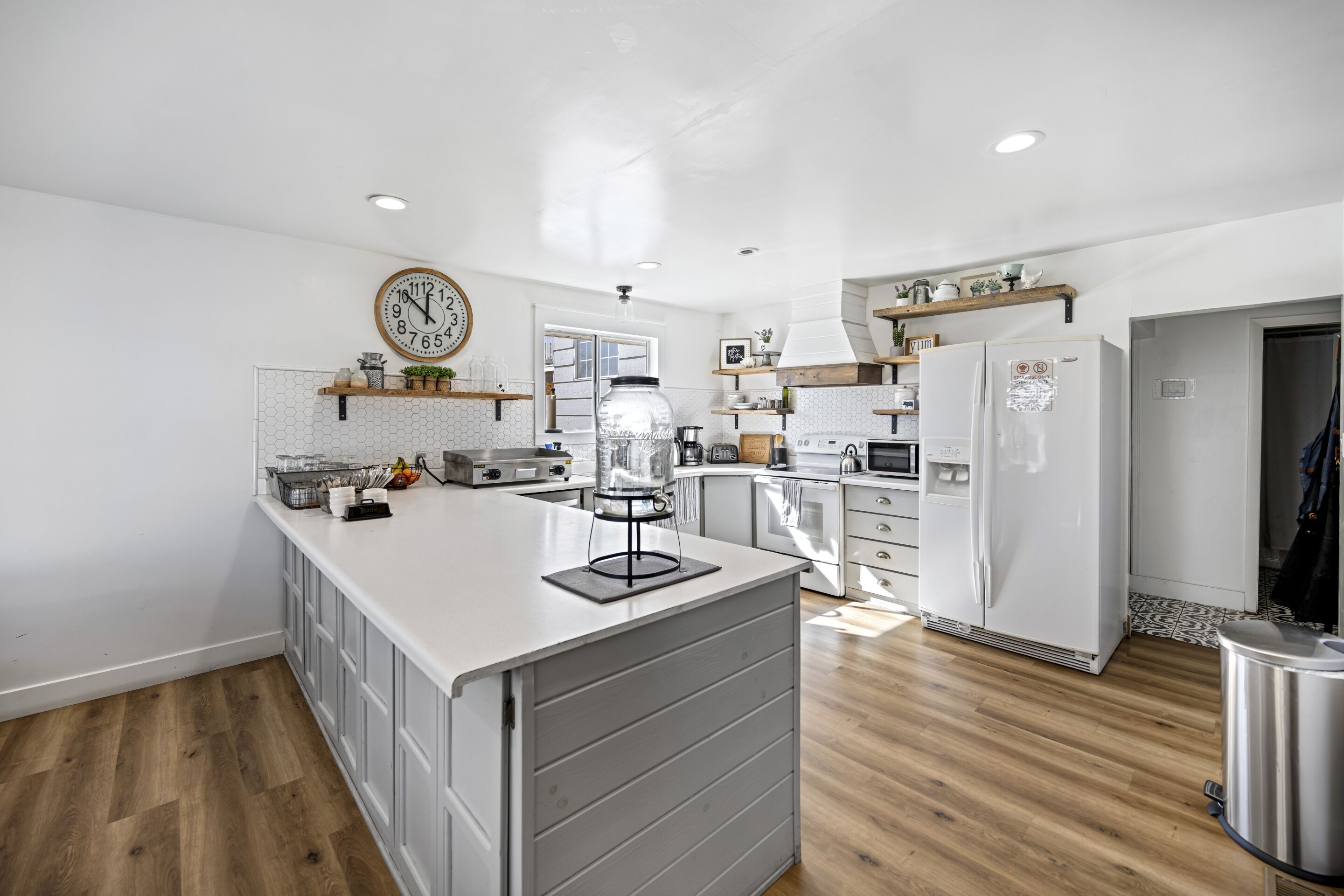 Une cuisine moderne avec des meubles blancs, un îlot central, une horloge et un plancher en bois.