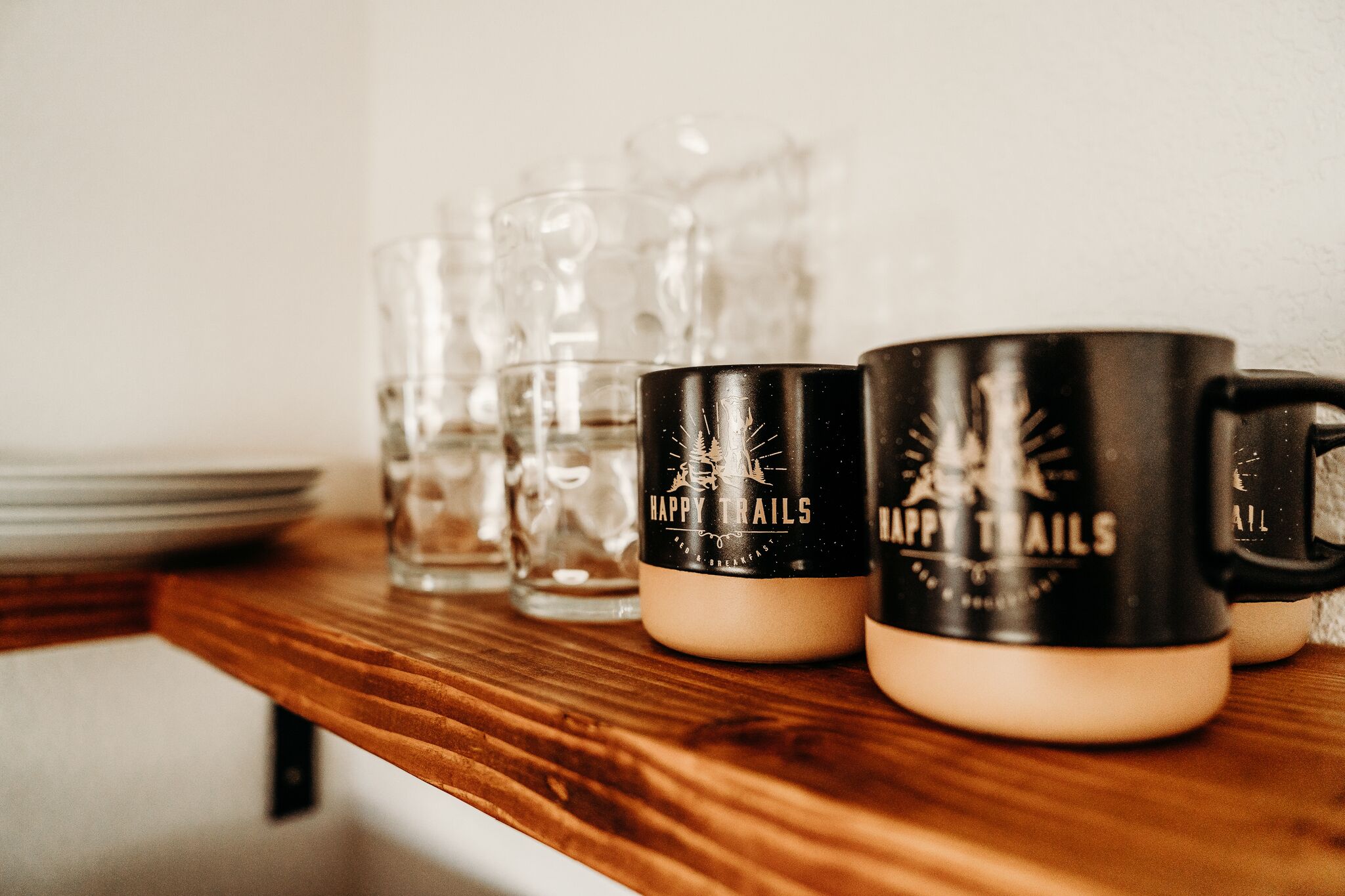Une étagère en bois avec des verres transparents et deux tasses noires ornées de l’inscription « HAPPY TRAILS ».