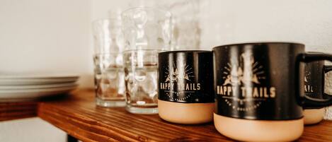 Una estantería de madera con vasos de vidrio transparente y dos tazas negras con el texto 'HAPPY TRAILS'.