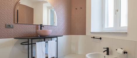 A modern bathroom with a round mirror, a sink with a vessel basin, a toilet, and a window.