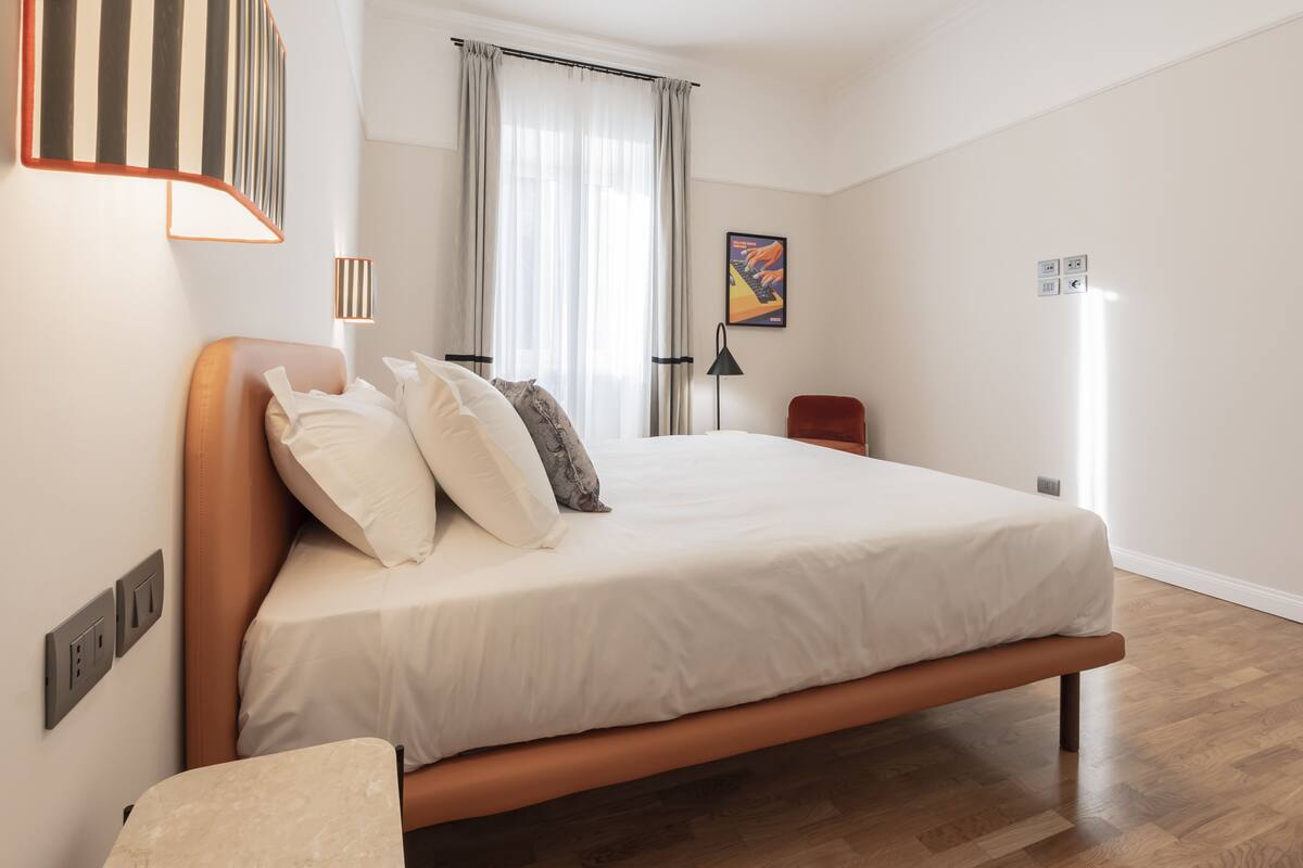 A modern bedroom with a wooden bed, white bedding, a bedside lamp, and a view of the outdoors through sheer curtains.
