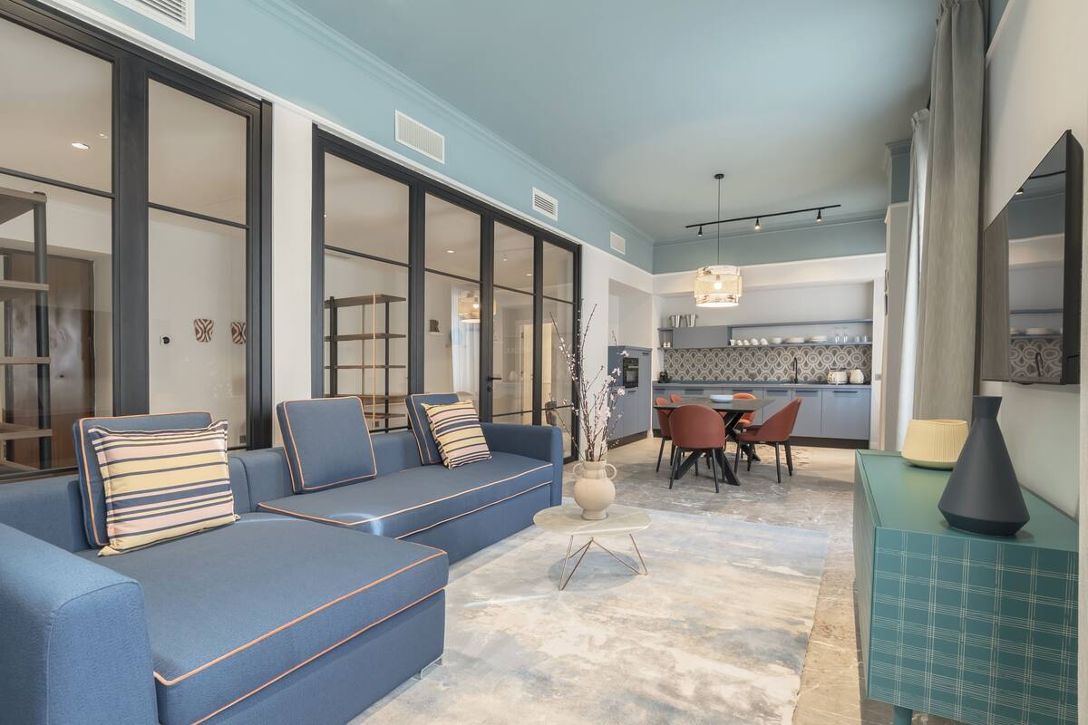 A modern living room with a blue sofa, a dining area with a table and chairs, and a kitchen in the background.