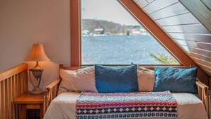 Un dormitorio acogedor con techo inclinado, cabecera de madera, lámpara de noche, cama con ropa de cama estampada y vista a un cuerpo de agua a través de un gran ventanal.