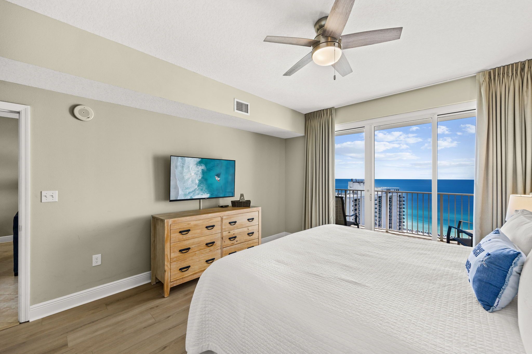 A hotel room with a large bed, a television, a wooden dresser, and a balcony with a view of the ocean.