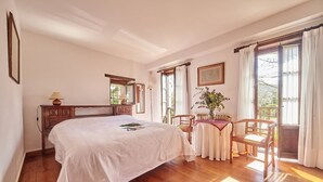 Un dormitorio con una cama grande, piso de madera, una mesita con sillas, una ventana con cortinas y un espejo.