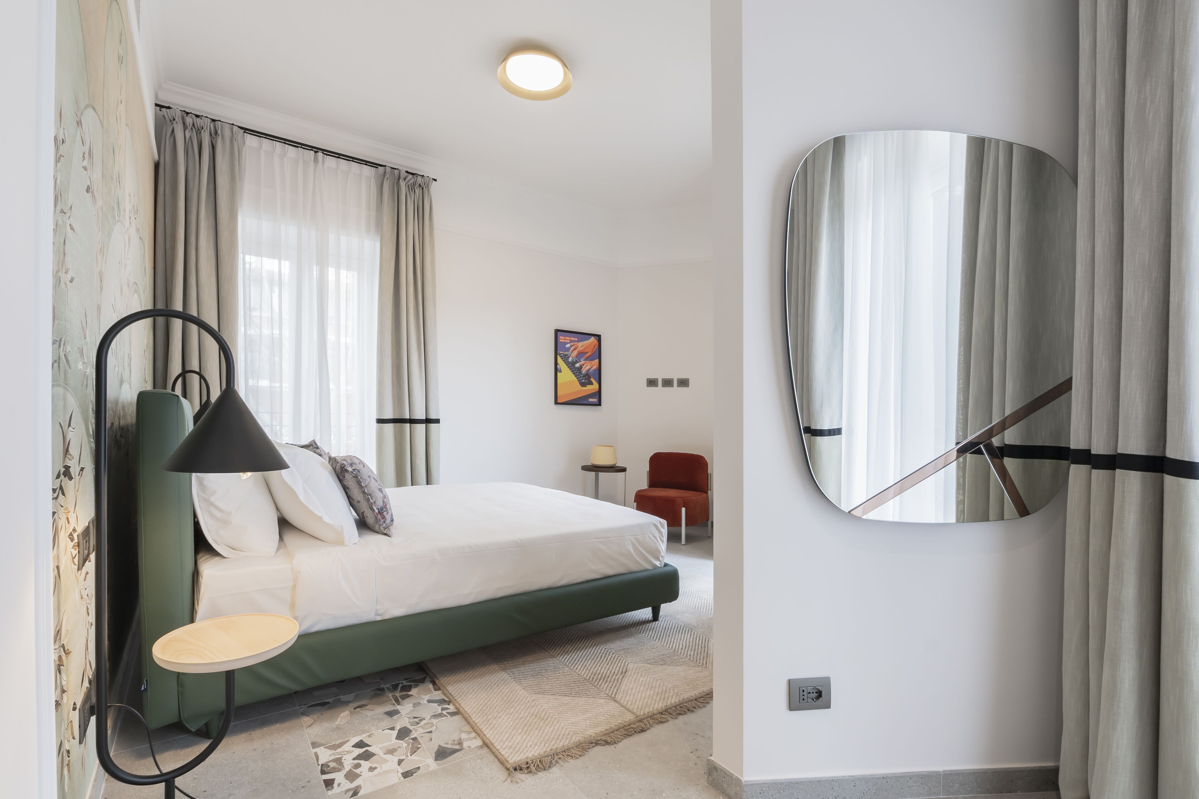 A bedroom with a bed, a bedside table, a lamp, a mirror, and a window with curtains.