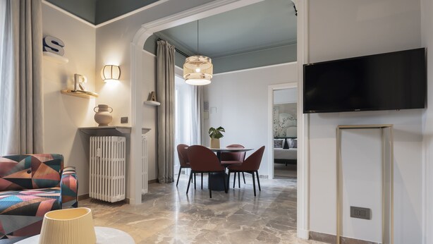 A modern living room with a sofa, a dining table with chairs, a TV mounted on the wall, and a lamp on a side table. - Be Mate Trastevere III (Rome)
