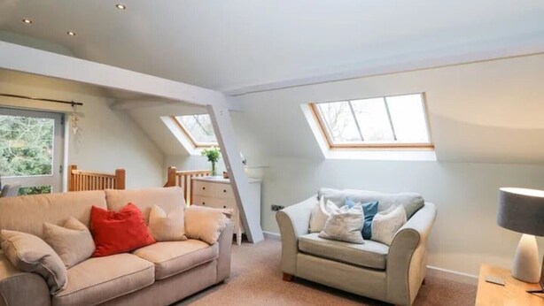 A spacious loft bedroom with a high ceiling, skylights, a large window, a sofa, a chair, and a bedside table.
