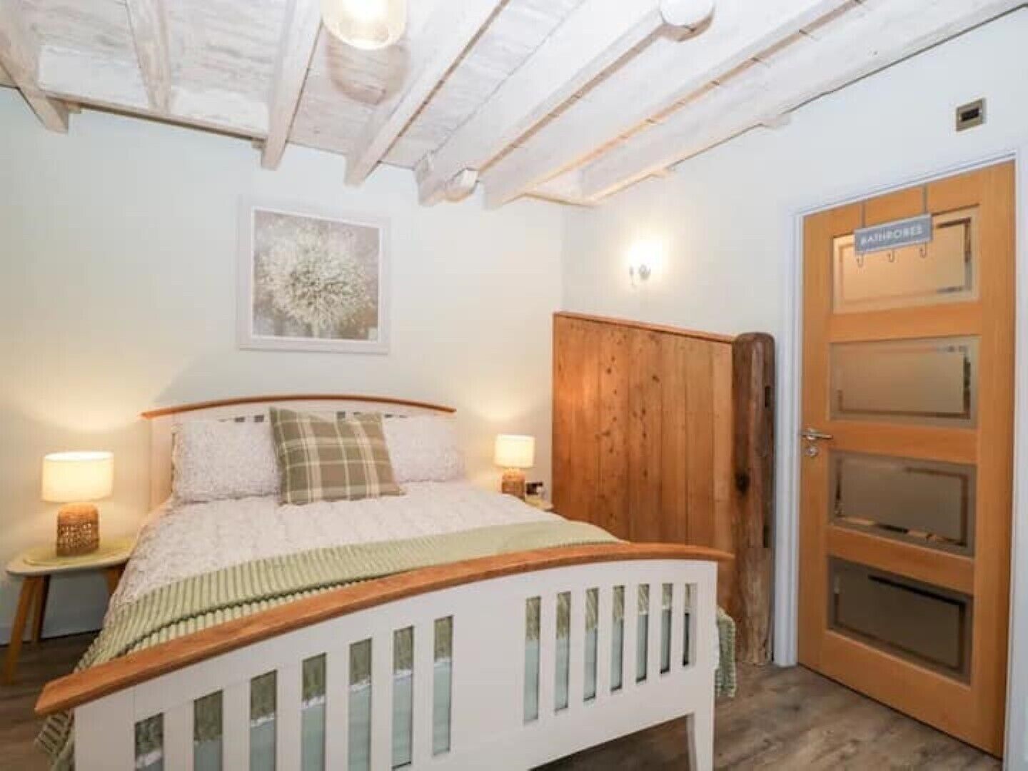 Un dormitorio con una cama de madera, una mesita de noche, una lámpara de pared, una puerta con un cartel y un techo con vigas a la vista.