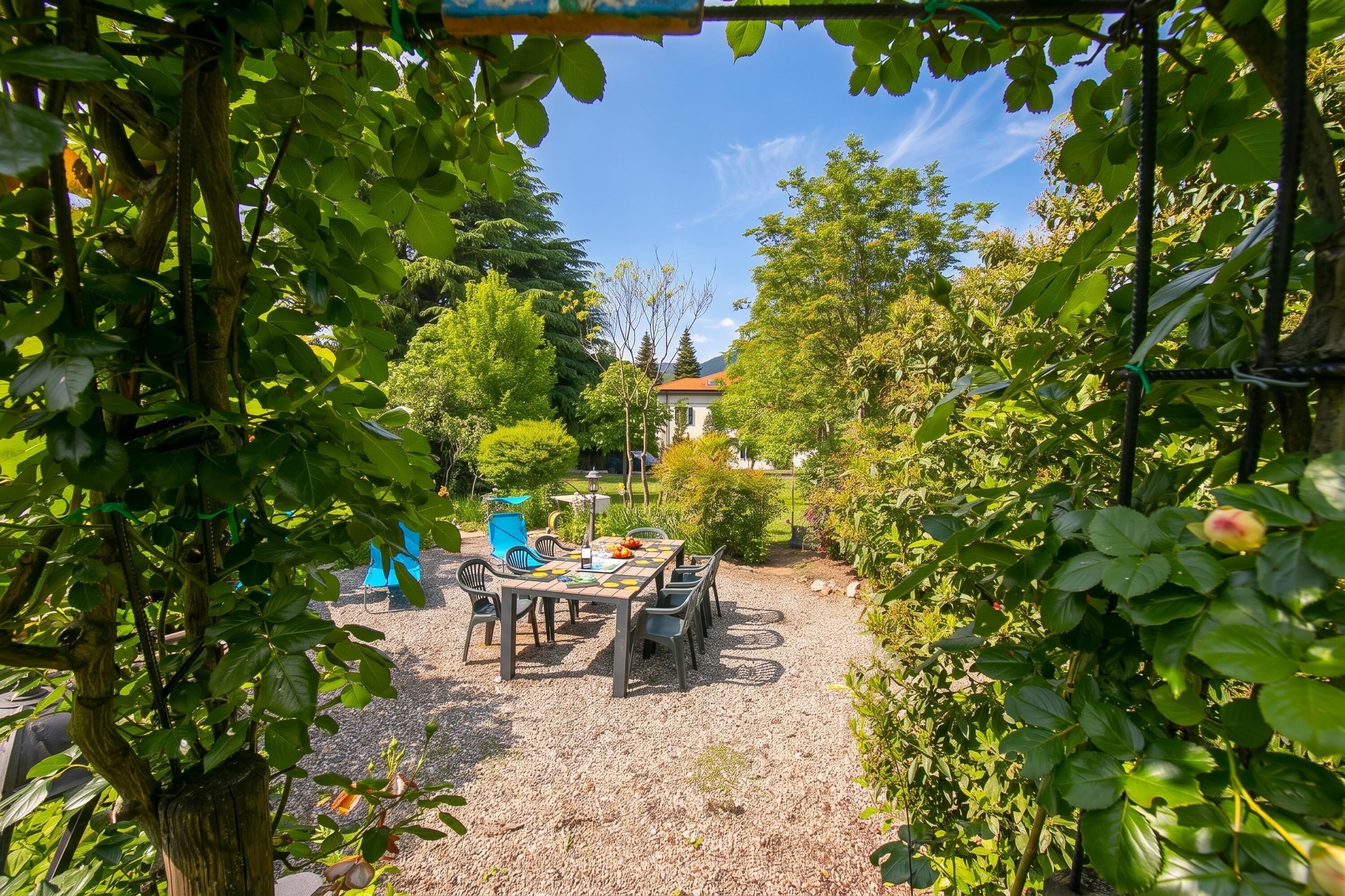 An outdoor dining area with a table and chairs, surrounded by greenery and a house in the background.