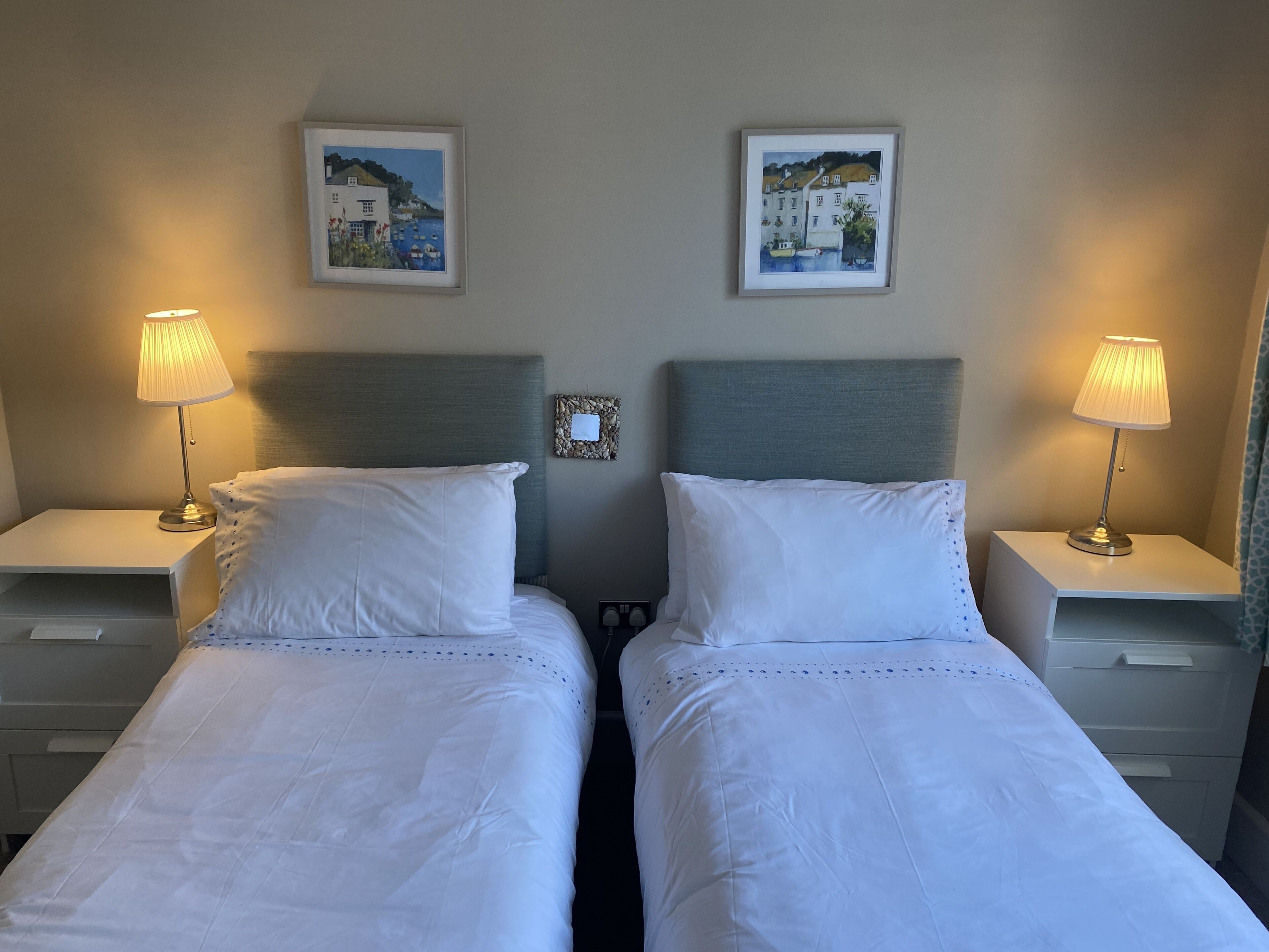 A hotel room with two single beds, each with a nightstand, a lamp, and a framed picture on the wall.