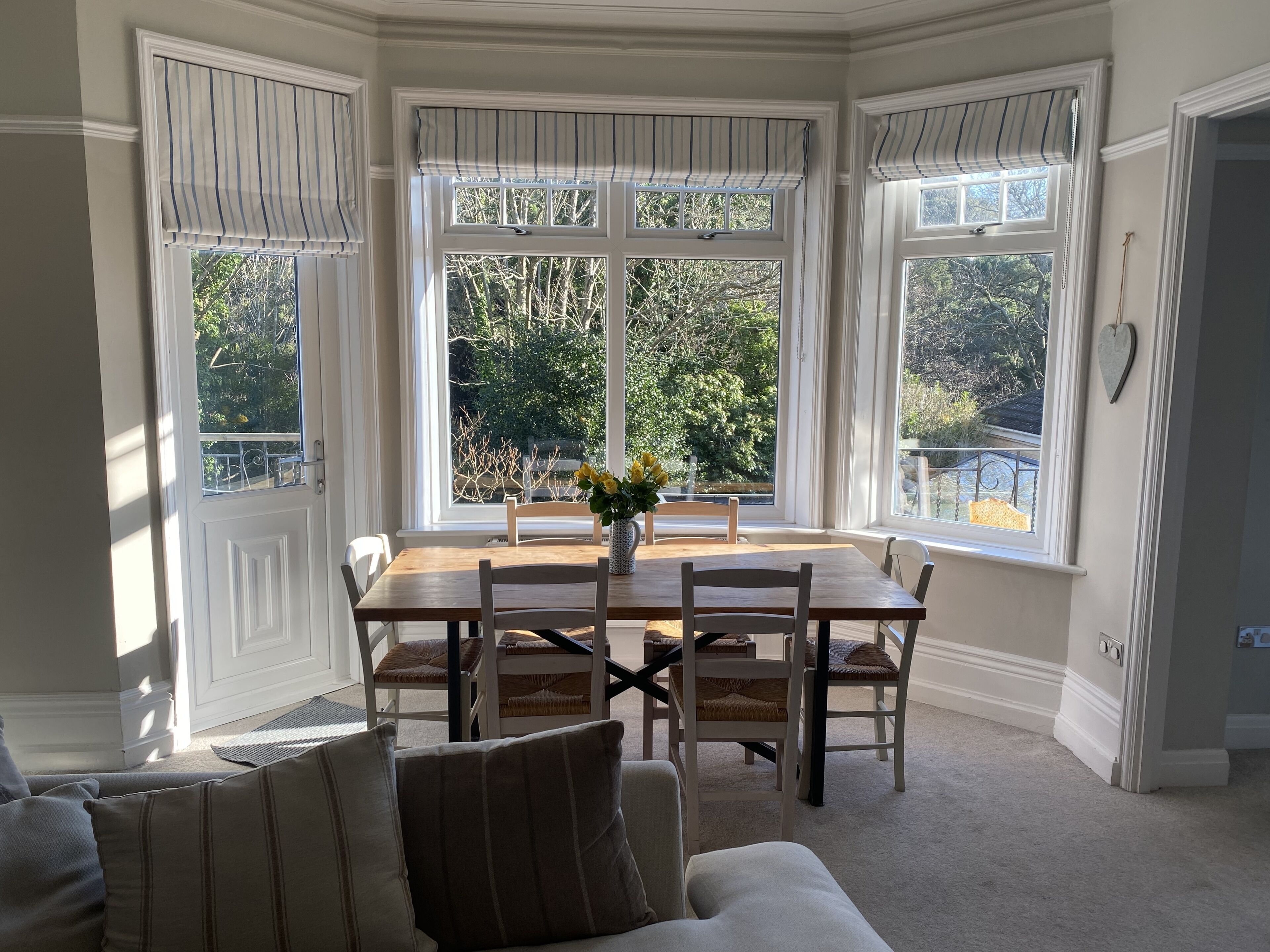 A dining area with a wooden table and chairs, large windows with white frames, and a view of greenery outside.