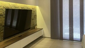 Un salon moderne avec un téléviseur à écran plat fixé sur un mur en pierre, une grande fenêtre avec des stores verticaux et un canapé.