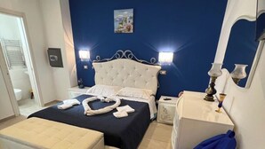 Une chambre à coucher avec un lit, des tables de chevet, une commode, un pouf bleu et une salle de bain visible à travers une porte ouverte.