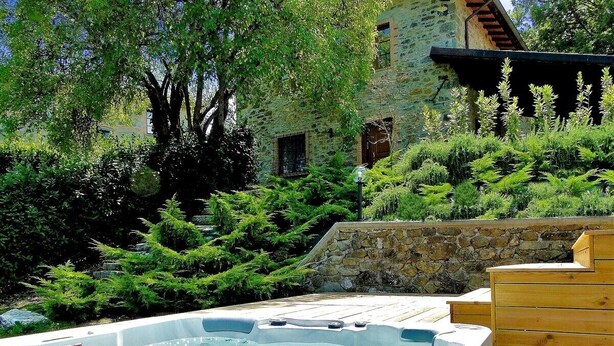 Un jacuzzi rodeado de vegetación exuberante y muros de piedra, con una terraza de madera rústica y un edificio de piedra al fondo.
