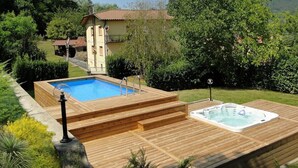 Una terraza de madera con un jacuzzi y una piscina, rodeada de follaje y una casa al fondo.