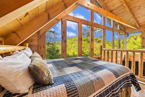 Une chambre dotée d’un grand lit, de poutres apparentes en bois au plafond et offrant une vue sur la verdure grâce à de grandes fenêtres.