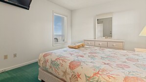 Une chambre à coucher comprenant un lit, une commode, un miroir, un téléviseur fixé au mur et une fenêtre avec des stores.