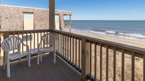 A wooden deck with two white plastic chairs, a beach view, and a building with a brick facade. - Oceanfront 3 Bedroom Condo at Golden Strand E3 (Kill Devil Hills)