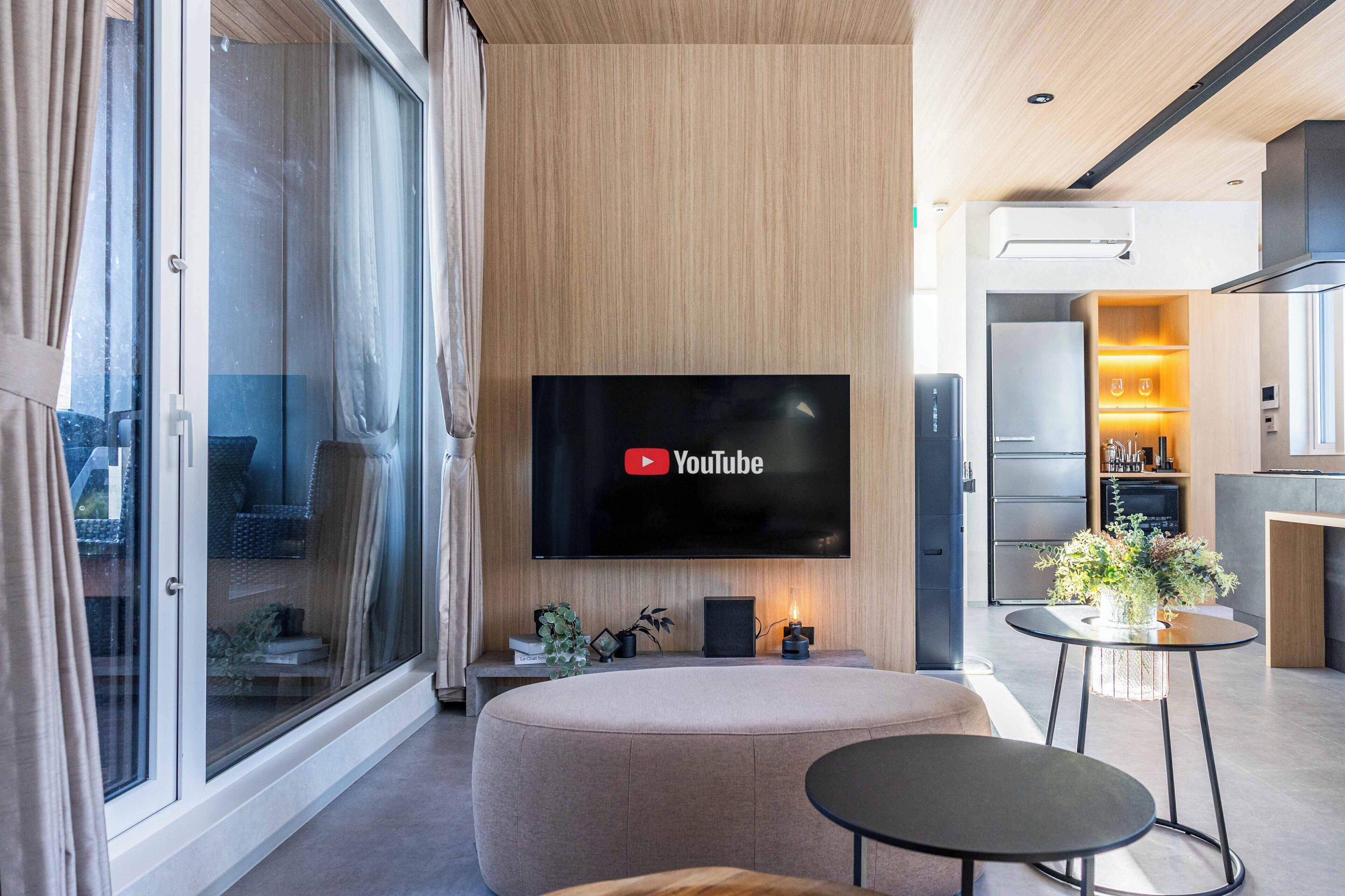A modern living room with a flat-screen TV mounted on the wall, a comfortable seating area, and a kitchen visible through an open door.