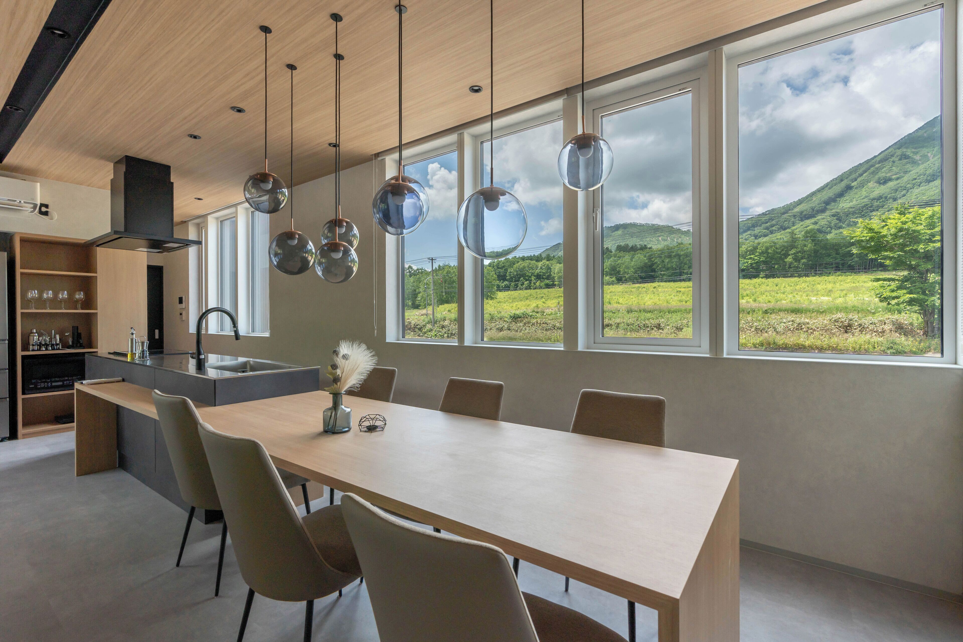 A modern kitchen with a large island, a dining area with a long table and chairs, and large windows offering a view of a green landscape.