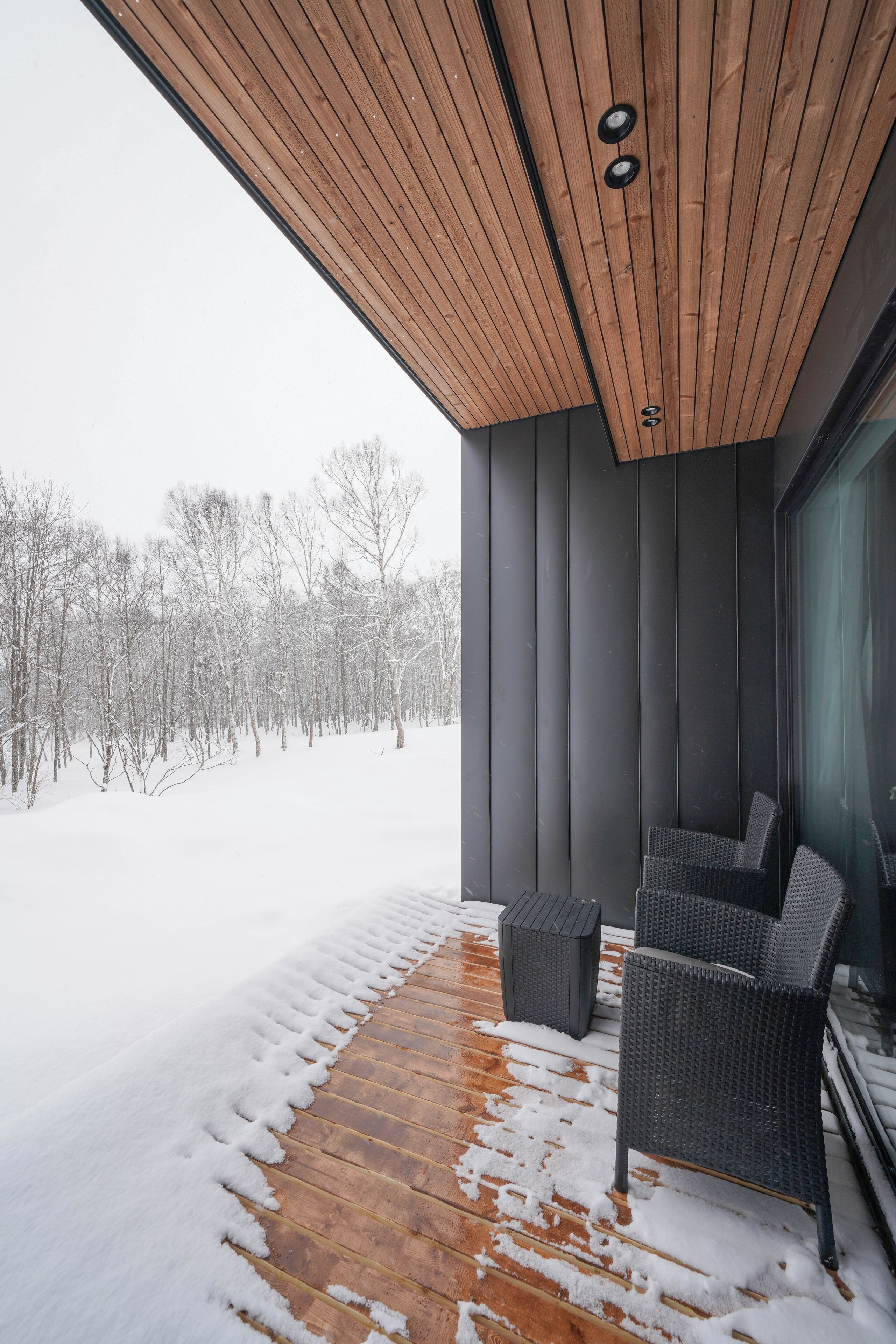 A modern exterior with a wooden ceiling, a glass door, and a snow-covered deck with two wicker chairs.