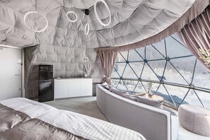 Un interior moderno con una cama grande, un sofá, una zona de cocina y vistas a través de un ventanal de diseño geométrico.