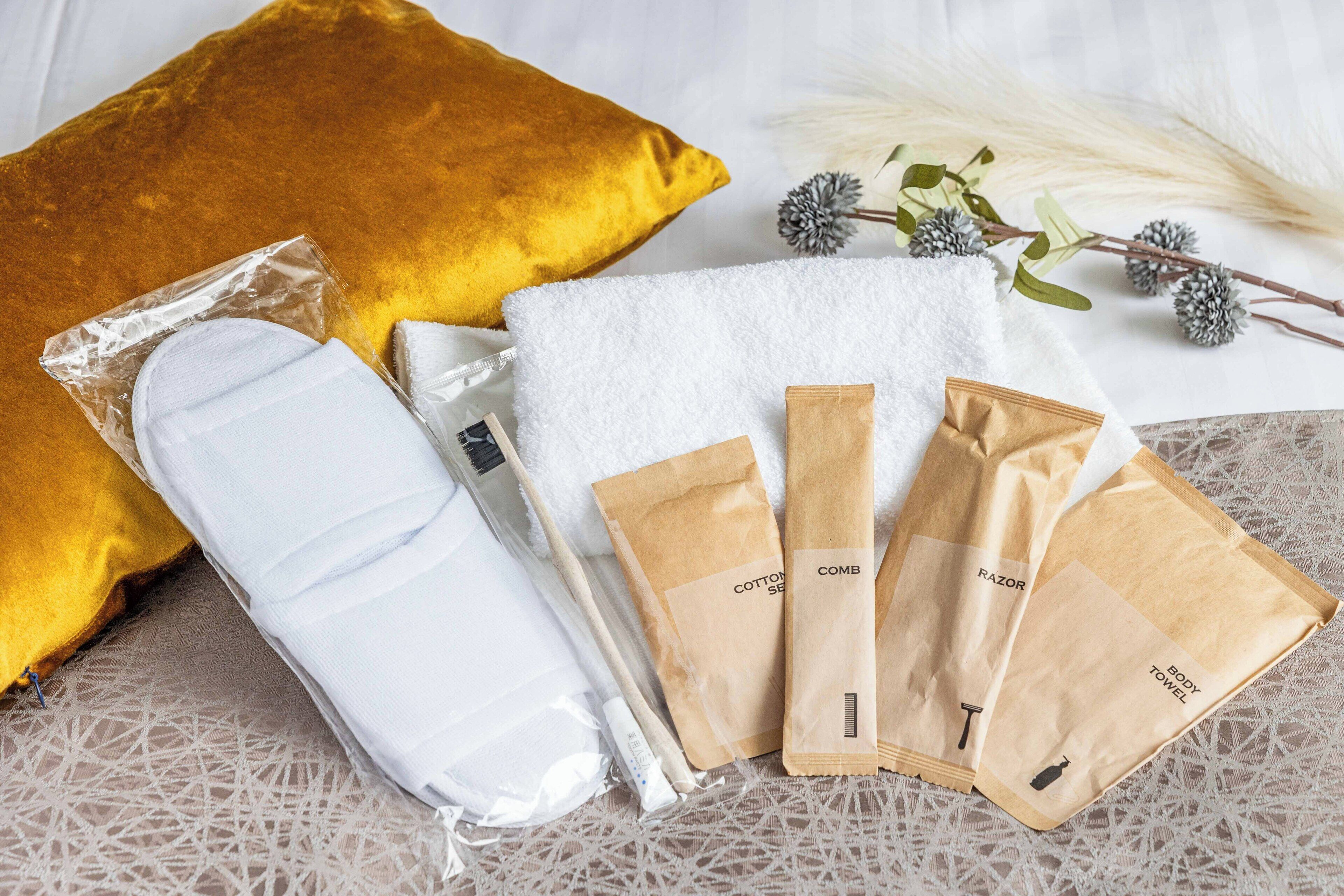 Ordentlich angeordnete Pflegeprodukte, darunter ein weißes Handtuch, ein gelbes Kissen und mehrere braune Papiertüten mit Kosmetikartikeln.