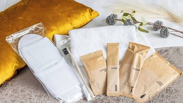 Ordentlich angeordnete Pflegeprodukte, darunter ein weißes Handtuch, ein gelbes Kissen und mehrere braune Papiertüten mit Kosmetikartikeln.