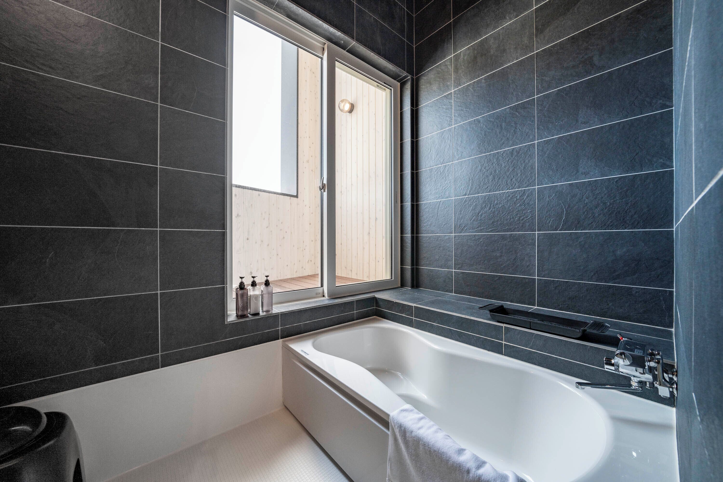 A modern bathroom with dark tiles, a large bathtub, a window, and a wooden floor.