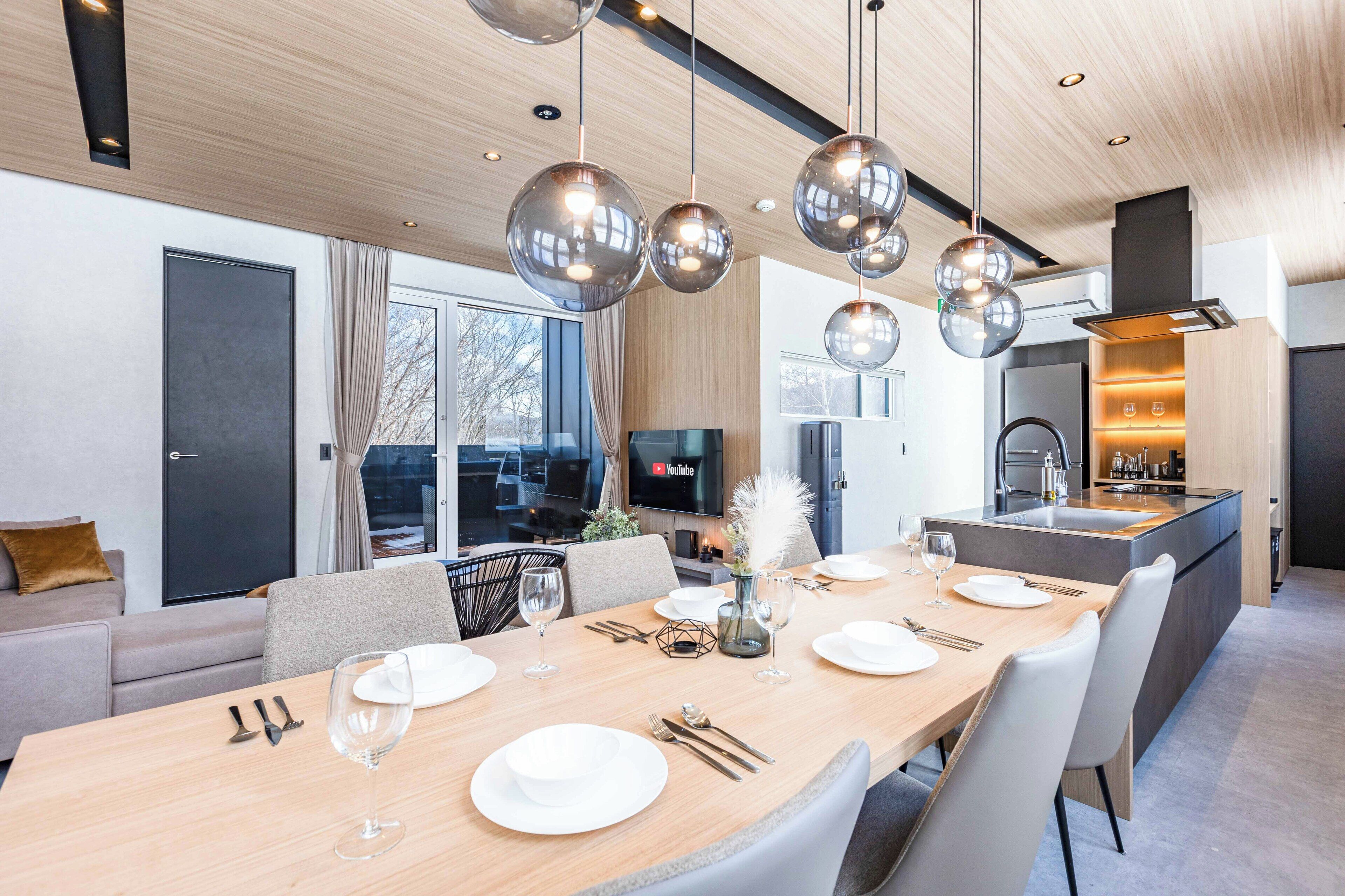Un comedor moderno con una mesa de madera preparada para seis comensales, una cocina al fondo y un ventanal con vistas al exterior.