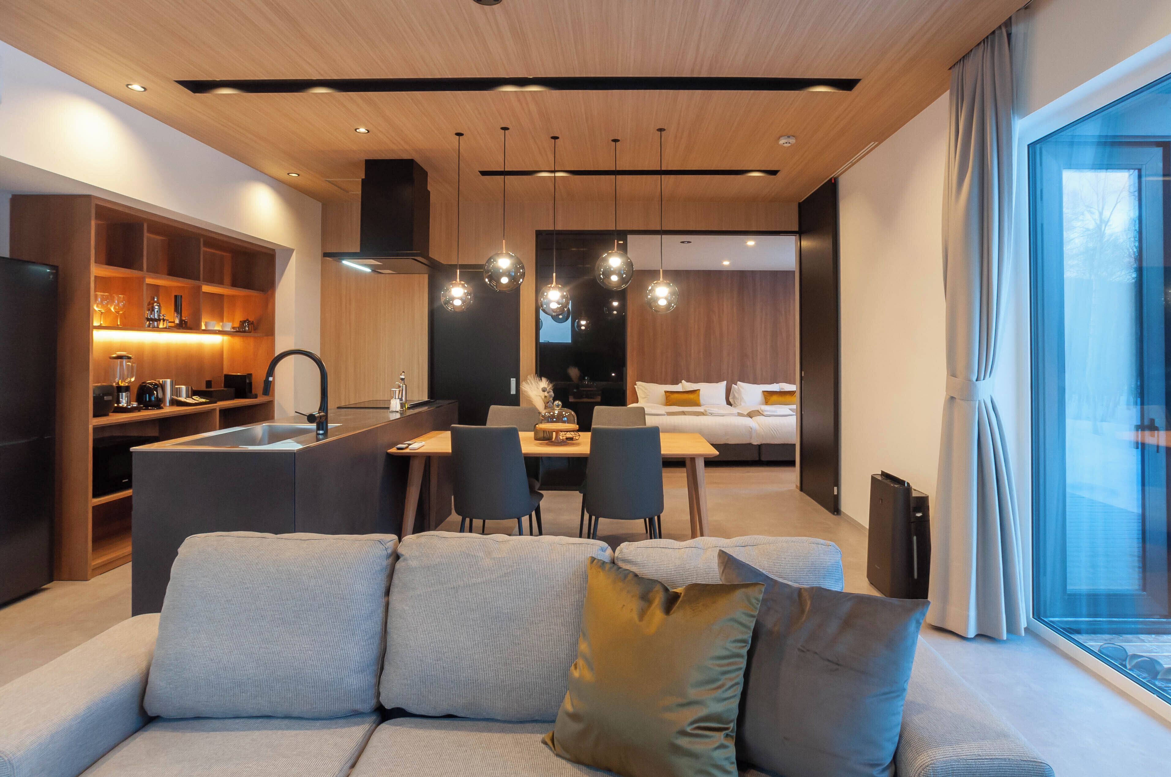 A modern hotel room with a kitchenette, a dining area, a sofa, and a large window.