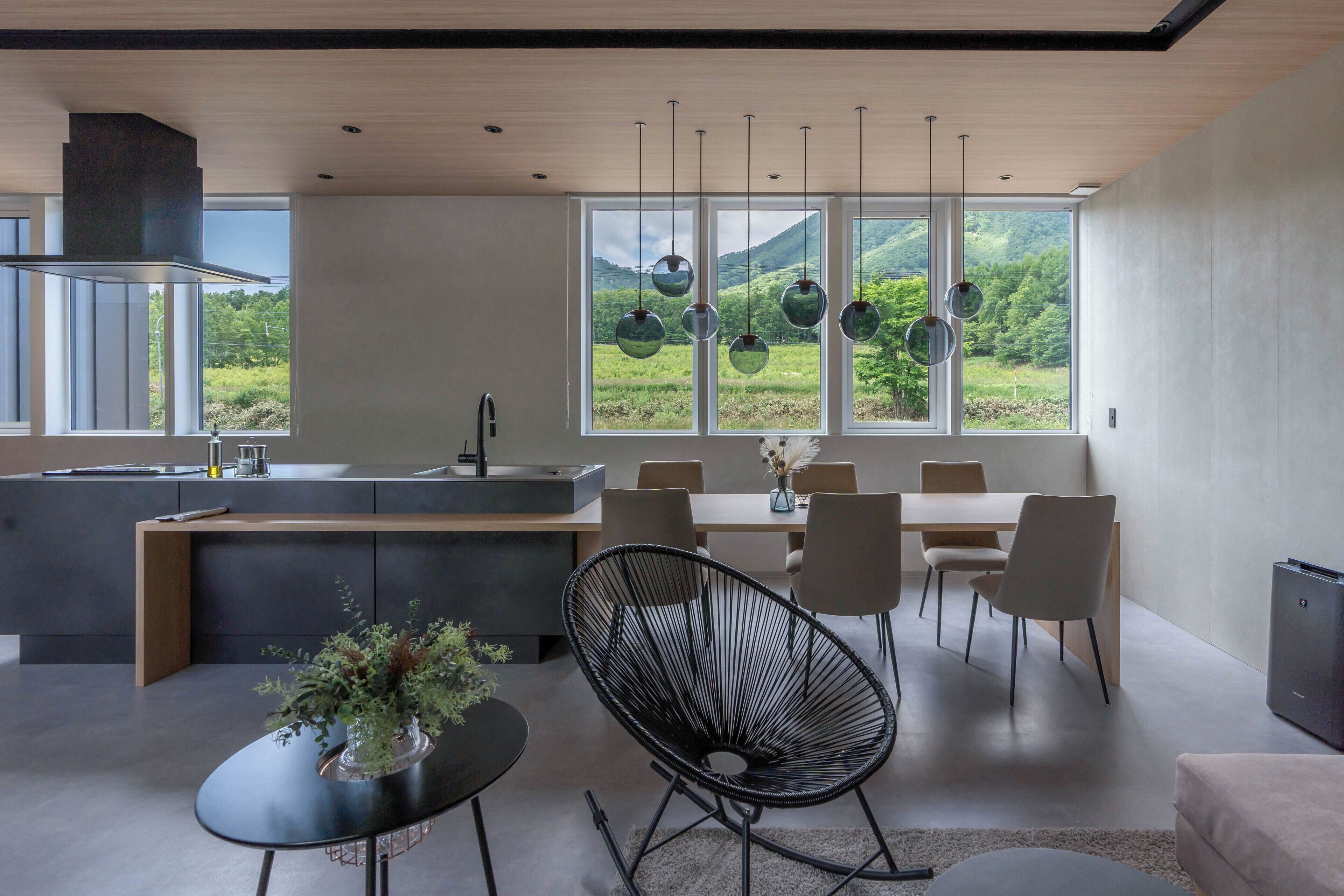 A modern kitchen with a central island, dining area with chairs, and large windows offering a view of greenery.
