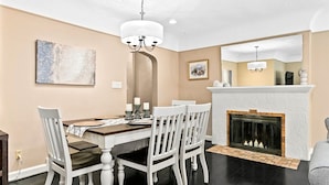Een eetkamer met een houten tafel en witte stoelen, een open haard en een kroonluchter.