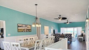 A spacious dining area with white chairs and a wooden table, a ceiling fan, and a view of the ocean through a sliding door. - Oceanfront House, Screen Porch, Boardwalk, Volleyball Sand Court, Game Room (Pawleys Island)