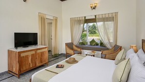Una habitación de hotel con una cama grande, un televisor en un mueble de madera, sillas de mimbre, una mesita y una ventana con cortinas.