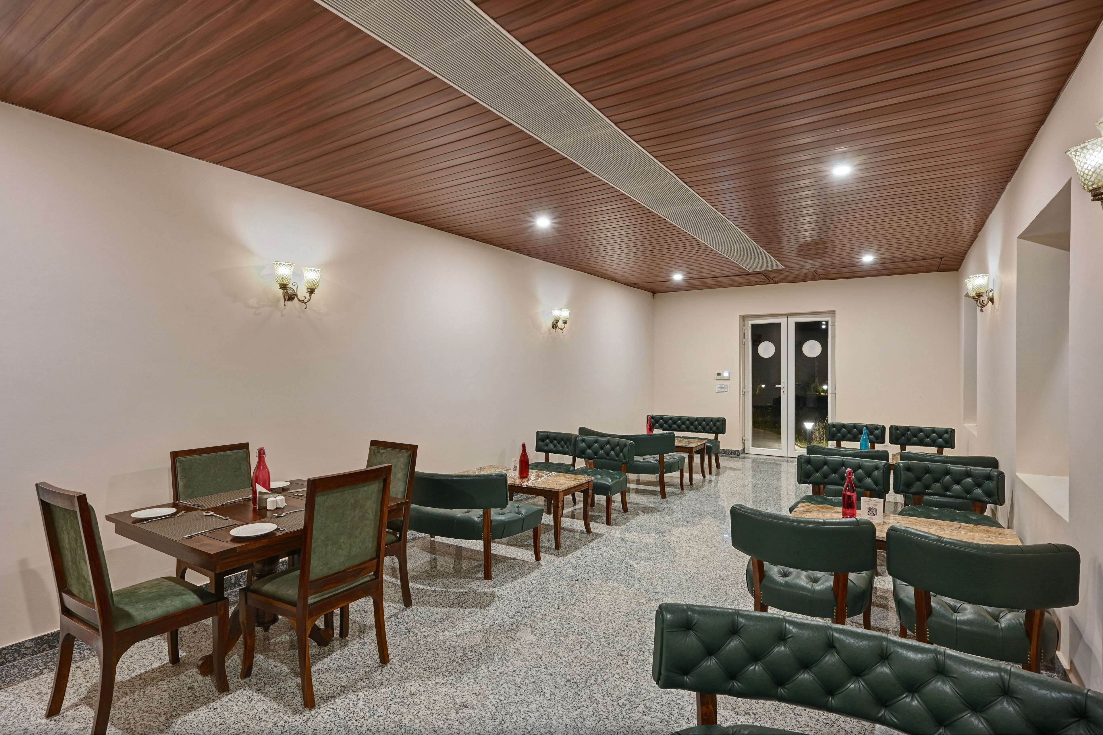 A dining area with wooden tables and green upholstered chairs, a wooden ceiling, and wall-mounted lighting.