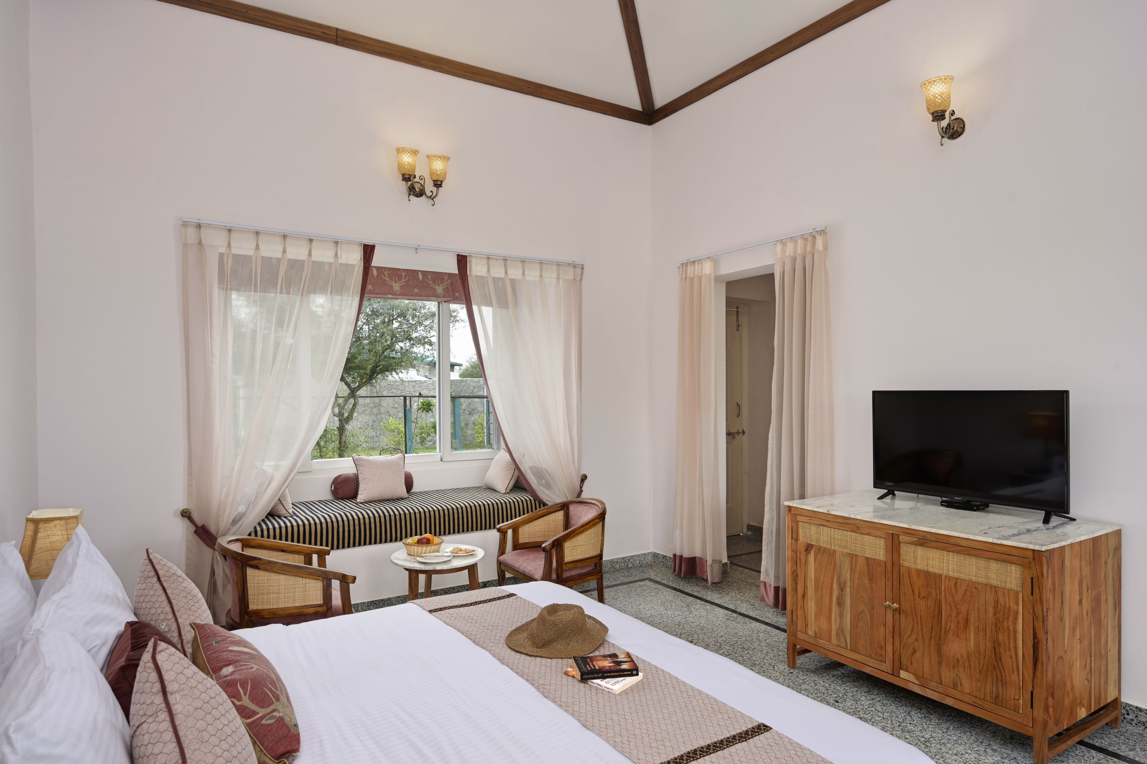 A hotel room with a bed, a television on a wooden cabinet, a seating area with a sofa and a small table, and a window with curtains.